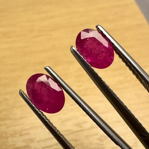 以下が含まれることがあります： 明るい木材の表面に、金属製のピンセットで保持された2つの楕円形にカットされた、ファセット加工されたルビーの宝石。 ルビーは深みのある鮮やかな赤色で、光を捉えるファセットが見えます。 ピンセットは銀色です。