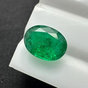 May include: Oval-cut emerald gemstone, displaying a vibrant green hue. The gem is set against a white surface, with facets catching the light. The gemstone is a focal point, showcasing its clarity and color.