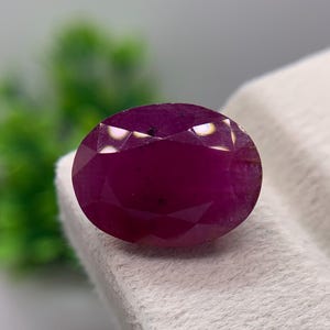 May include: A deep magenta-colored oval gemstone with a faceted surface. The stone features a polished finish with multiple triangular and diamond-shaped facets that reflect light, creating a brilliant appearance across its rounded, symmetrical shape.