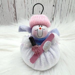 May include: A snowman ornament wearing a pink knit hat and a light blue scarf. The snowman holds a pink nail file, a small bottle of pink glitter nail polish, and a single silver glitter artificial fingernail. A black metal hook is attached to the top.