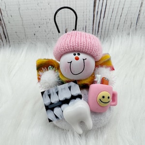 May include: A dental-themed ornament with a smiling face wearing a pink knitted hat. It features a dental X-ray, a tooth, and a pink mug with a smiley face. The ornament has a black loop for hanging.