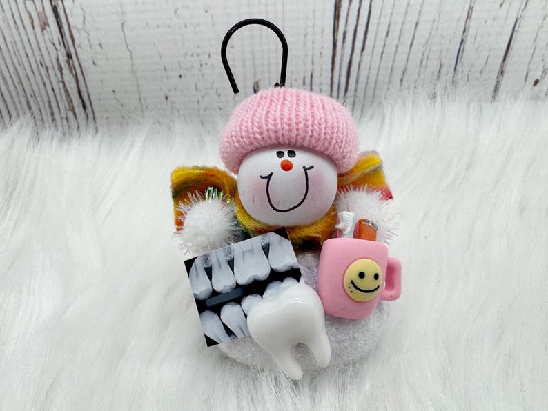 May include: A dental-themed ornament with a smiling face wearing a pink knitted hat. It features a dental X-ray, a tooth, and a pink mug with a smiley face. The ornament has a black loop for hanging.