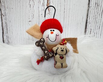 May include: A snowman ornament featuring a white round body, a red knit hat, and a tan felt scarf. The snowman holds a dark brown bone-shaped treat with gold heart accents and a small tan dog figurine with a red heart on its chest.
