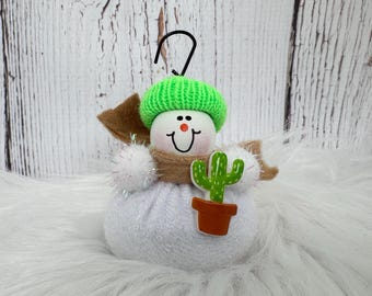 May include: A white snowman ornament with a bright green knit hat, a tan felt scarf, and a small brown potted cactus decoration. The snowman has a round white head with a painted face, a black metal hanging hook, and a soft white fabric body.