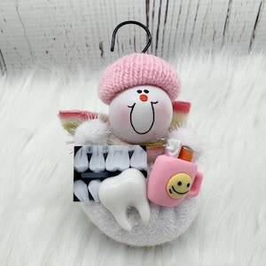 May include: A dental-themed ornament with a smiling face wearing a pink knitted hat. It features a tooth, dental X-ray, and a pink mug with a smiley face. The ornament has a black hook for hanging.