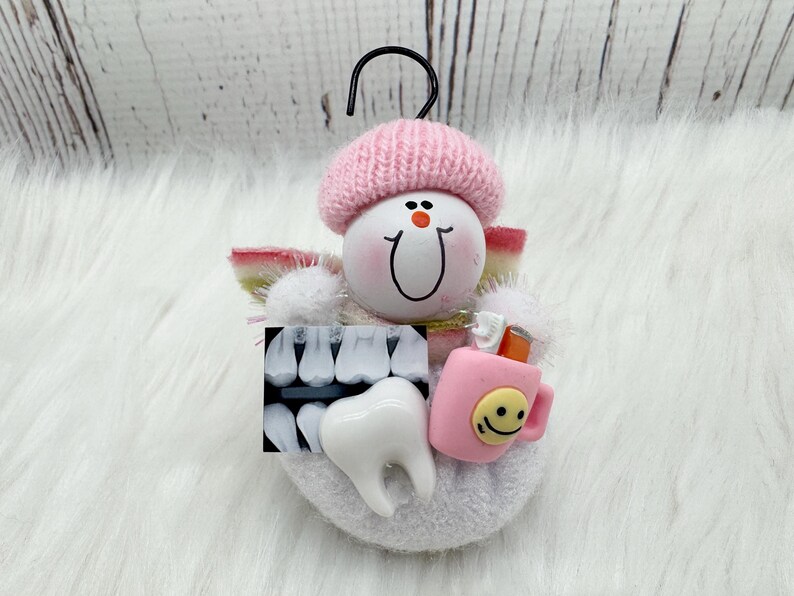 May include: A dental-themed ornament with a smiling face wearing a pink knitted hat. It features a tooth, dental X-ray, and a pink mug with a smiley face. The ornament has a black hook for hanging.