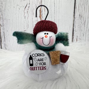May include: A plush snowman ornament wearing a dark red knit hat and a dark green scarf. It features a round white body, a painted face with an orange nose, and a circular badge that reads CORKS ARE FOR QUITTERS with a wine bottle and cork icons.