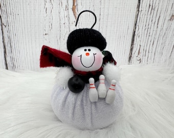 May include: A round white fabric snowman ornament featuring a black knit hat, a red and black plaid scarf, and a black metal hanging hook. The snowman holds a small black bowling ball and three white bowling pins with red stripes on its front.