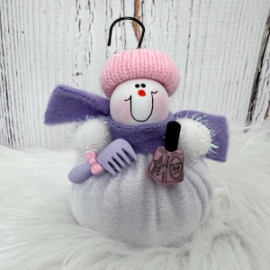 May include: A snowman ornament featuring a white round body, a pink knit hat, and a purple felt scarf. The snowman holds a small lavender hair comb with a pink bow and a miniature bottle of pink nail polish with a black cap and star graphic.
