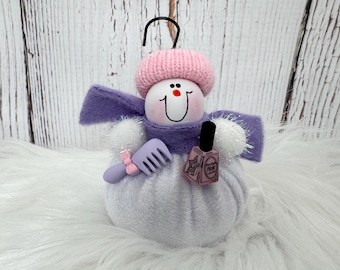 May include: A snowman ornament featuring a white round body, a pink knit hat, and a purple felt scarf. The snowman holds a small lavender hair comb with a pink bow and a miniature bottle of pink nail polish with a black cap and star graphic.