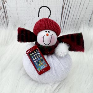 May include: A snowman ornament featuring a white fabric body, a red knit hat, and a dark red plaid scarf. The snowman has a painted face with a small orange nose and holds a miniature red smartphone with a colorful app icon display on its screen.