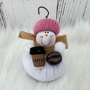 May include: A snowman decoration with a pink knitted hat, holding a coffee cup and a coffee bean. The snowman has a painted face with a smiling expression and a carrot nose. The ornament has a black wire loop for hanging.