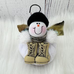 May include: A snowman ornament with a black hat, a smiling face, and tan combat boots. The snowman has white fluffy arms and wings with a touch of glitter. A black metal hook is attached to the top for hanging.