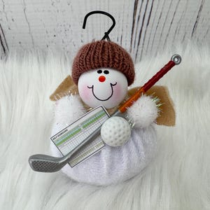 May include: A snowman-shaped Christmas ornament with a brown knitted hat, holding a golf club, scorecard, and golf ball. The ornament has a smiling face with rosy cheeks and a black hook for hanging. The wings are beige.