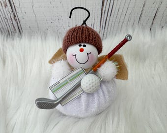 May include: A snowman-shaped Christmas ornament with a brown knitted hat, holding a golf club, scorecard, and golf ball. The ornament has a smiling face with rosy cheeks and a black hook for hanging. The wings are beige.