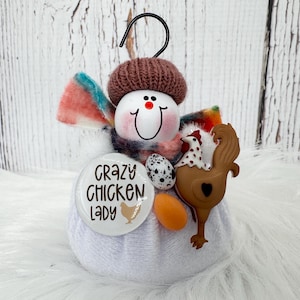 May include: A white fabric ornament featuring a snowman head with a brown knit hat, a colourful scarf, and a black hanging hook. It includes a round button with the text Crazy Chicken Lady, a small speckled egg, an orange egg, and a brown rooster.