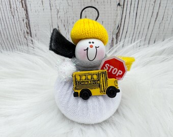 May include: A charming ornament depicting a snowman-like character with a yellow hat and black wings. It holds a yellow school bus and a red stop sign. The ornament is made of various materials and has a loop for hanging.