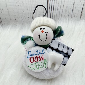 May include: A snowman ornament with a white knitted hat and a smiling face. It features a "Dental Crew" button, a tooth model, and a dental X-ray image. The ornament has a black hook for hanging.