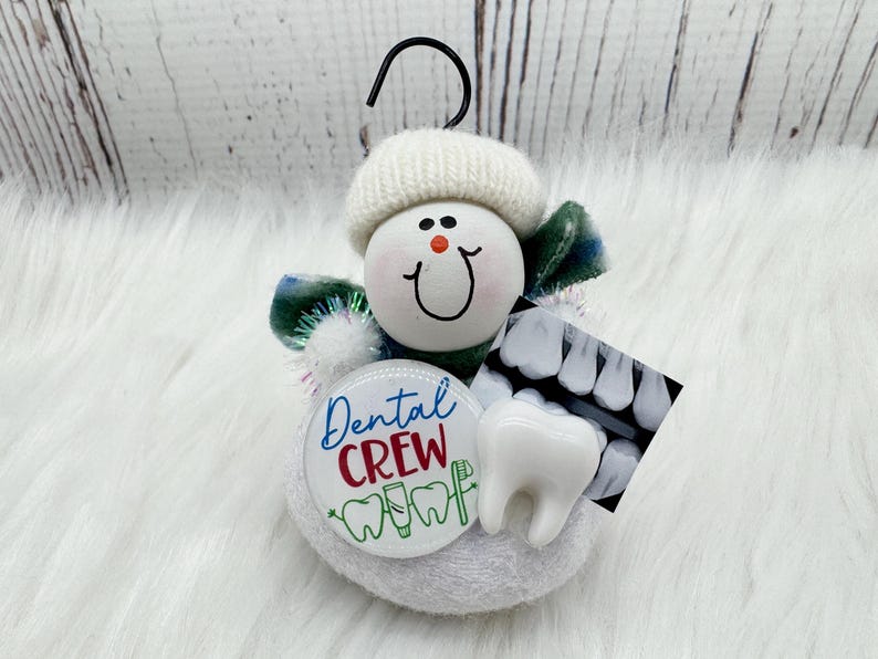 May include: A snowman ornament with a white knitted hat and a smiling face. It features a "Dental Crew" button, a tooth model, and a dental X-ray image. The ornament has a black hook for hanging.