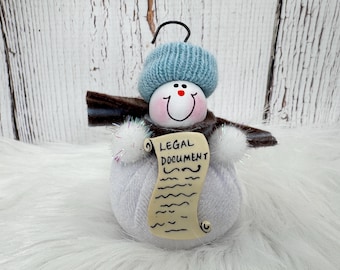 May include: A snowman ornament made of white yarn with a light blue knit hat, a brown felt scarf, and white pom-pom hands. The snowman holds a beige scroll with the text LEGAL DOCUMENT printed in black, and features a black wire hanging loop.