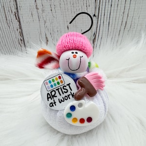 May include: A snowman-shaped ornament with a pink knit hat and a colorful scarf. It features a smiling face, a paintbrush, a palette, and a button that says "ARTIST at work". The ornament has a black loop for hanging.