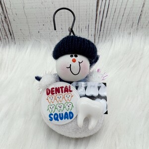 May include: A snowman-style ornament with a black hat and a smiling face. It features a "Dental Squad" button with cartoon teeth, a tooth model, and a dental X-ray image. The ornament is designed to hang.