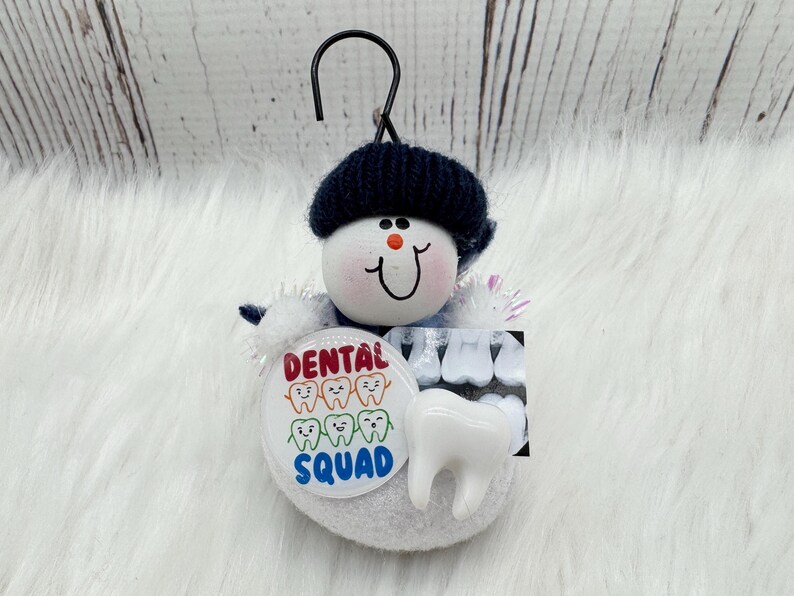 May include: A snowman-style ornament with a black hat and a smiling face. It features a "Dental Squad" button with cartoon teeth, a tooth model, and a dental X-ray image. The ornament is designed to hang.