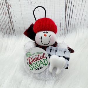 May include: A snowman decoration with a red knitted hat and scarf. It features a Dental Squad badge, a tooth charm, and a dental X-ray image. The ornament has a black loop for hanging.