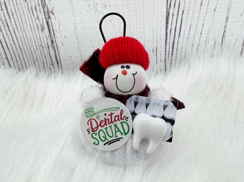 May include: A snowman decoration with a red knitted hat and scarf. It features a Dental Squad badge, a tooth charm, and a dental X-ray image. The ornament has a black loop for hanging.