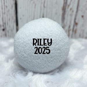 May include: A white, spherical object with the name "RILEY" and the year "2025" printed in black. The object is made of a soft, textured material and rests on a fluffy white surface.