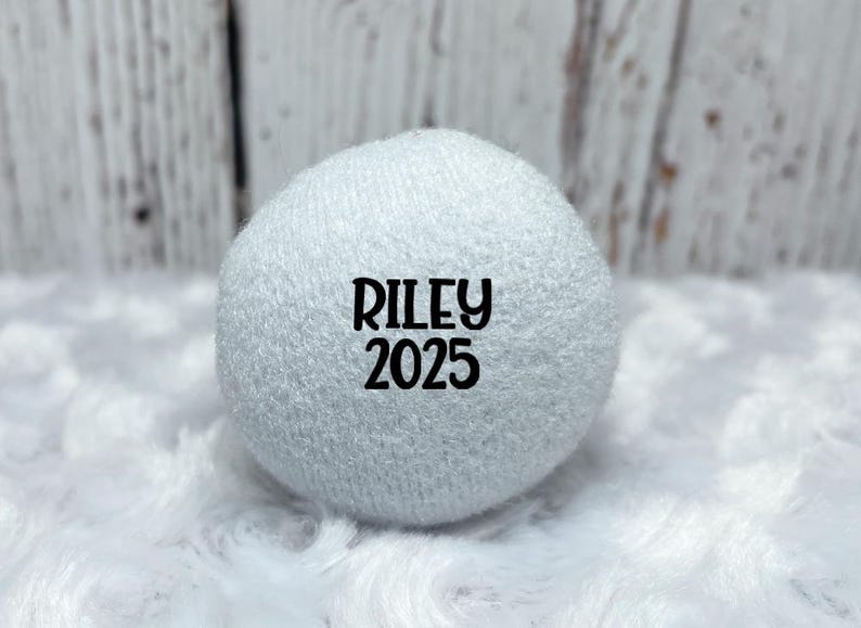 May include: A white, spherical object with the name "RILEY" and the year "2025" printed in black. The object is made of a soft, textured material and rests on a fluffy white surface.