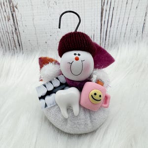 May include: A dental-themed ornament depicting a snowman with a burgundy hat and scarf. It holds a tooth, a pink mug with a smiley face, and a dental x-ray. The ornament has a black hook for hanging. The snowman's face has a smiling expression.