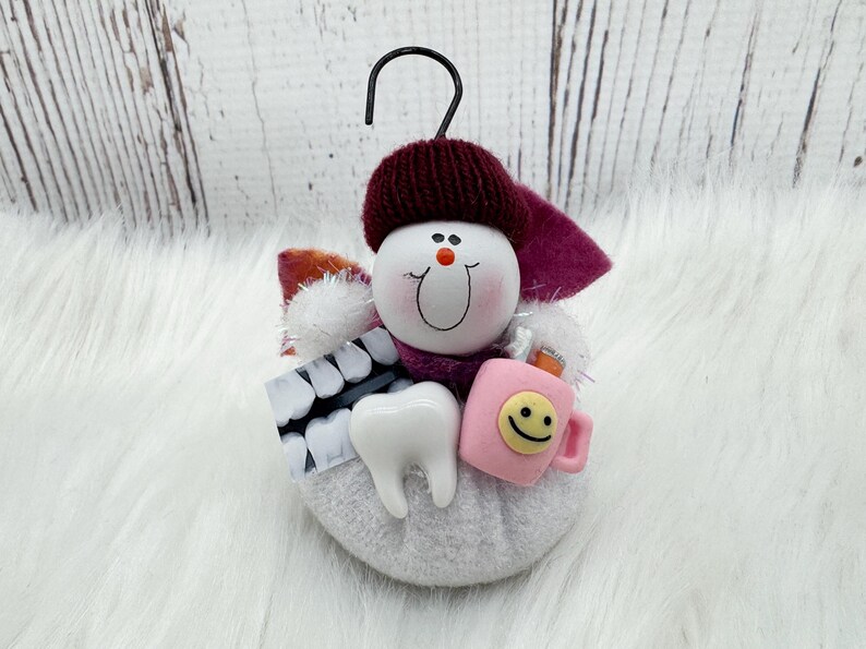 May include: A dental-themed ornament depicting a snowman with a burgundy hat and scarf. It holds a tooth, a pink mug with a smiley face, and a dental x-ray. The ornament has a black hook for hanging. The snowman's face has a smiling expression.