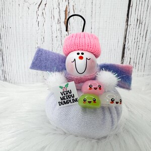 May include: A snowman ornament featuring a white fabric body, a pink knit hat, and a purple and blue scarf. It holds three small, colorful, smiling dumpling charms and a square sign that reads VERY MERRY DUMPLING with a small holly illustration.