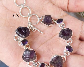 May include: A silver bracelet with raw, dark purple amethyst stones. The bracelet has a toggle clasp and is made up of various sized amethyst pieces. The letters "CSJ" are visible on the image.