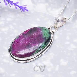Puede incluir: Un colgante de forma ovalada con una piedra preciosa de rubí zoisita pulida con inclusiones de color magenta vibrante y verde intenso, engastada en un bisel de plata liso con un borde ranurado decorativo, suspendido de una cadena de plata, con las letras CSJ.