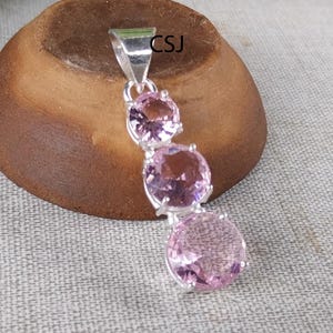 May include: A silver pendant featuring three round, light pink gemstones. The gemstones are set in a silver setting and are connected in a vertical line. The pendant has a loop at the top for a chain. The letters "CSJ" are visible on the top of the pendant.