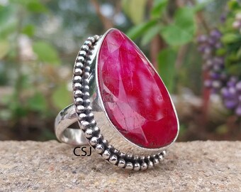 May include: A silver ring with a large, faceted, teardrop-shaped ruby gemstone. The ruby is a vibrant red colour. The ring has a beaded border and the initials "CSJ" are visible on the band.