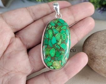Puede incluir: Colgante ovalado con una vibrante piedra verde y marrón engastada en un marco de metal plateado. La piedra tiene un aspecto de mosaico con diferentes tonos de verde. El colgante tiene una fianza plateada para sujetar a una cadena.