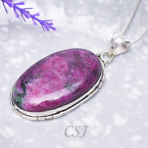 Puede incluir: Un colgante de forma ovalada con una piedra preciosa pulida de color magenta y negro engastada en un bisel de plata, suspendido de una cadena de plata. La piedra muestra una textura moteada con inclusiones rosa oscuro y verde oscuro. Las letras CSJ son visibles.