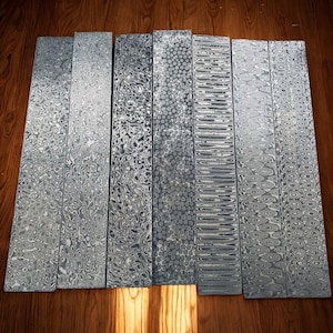 Può includere: Una collezione di otto lastre metalliche rettangolari con intricati motivi a vortice in tonalità di grigio e argento. I motivi variano, mostrando una gamma di trame e design. Le lastre sono disposte su una superficie di legno.
