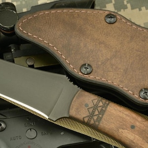 Puede incluir: Un cuchillo negro de hoja fija con mango de madera marrón y funda de cuero marrón. La hoja del cuchillo tiene un borde plateado. El mango tiene un patrón geométrico decorativo. La funda está asegurada con tornillos negros.