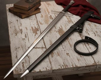 May include: A long, silver sword with a black handle and crossguard rests on a distressed white wooden table. A black scabbard and belt are next to the sword. A stack of old books and a lit candle are in the background.