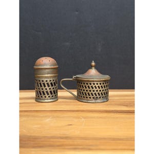 May include: Two antique metal containers. One is a salt shaker with a perforated design and a domed top. The other is a small lidded cup with a handle, also featuring a perforated design. Both have a tarnished, aged appearance.