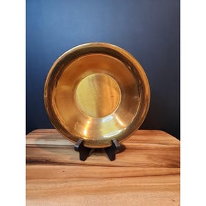 Puede incluir: Un cuenco redondo de metal dorado con un borde ancho. El cuenco se muestra sobre un pequeño soporte negro, sobre un fondo azul oscuro. El interior del cuenco tiene una superficie lisa y reflectante.