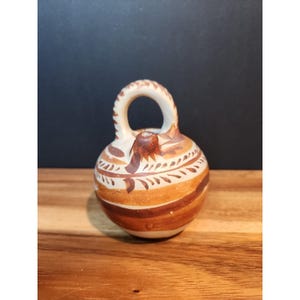 Puede incluir: Jarra de cerámica pequeña y redonda con asa y pico. La jarra está decorada con bandas marrones y beige y patrones en forma de hojas. El asa tiene forma de bucle. La jarra está sobre una superficie de madera.