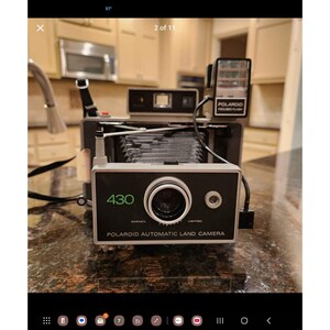 Puede incluir: Una cámara Polaroid Automatic Land Camera modelo 430 vintage, con un cuerpo negro y plateado. La cámara tiene una lente grande, un accesorio de flash y el texto "430" y "Polaroid Automatic Land Camera" en la parte delantera. Un clásico.