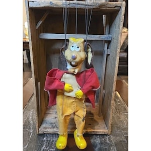 Può includere: Un burattino vintage di un personaggio dei cartoni animati con un viso abbronzato, orecchie marroni e una corona gialla. Il burattino indossa una giacca rossa, pantaloni gialli e scarpe gialle. È sospeso da fili ed esposto in una scatola di legno.