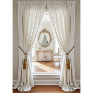 May include: Cream-colored curtains framing a view of a room with a decorative mirror and antique furniture. The curtains are held back with gold-colored rope and tassels, creating an elegant and inviting atmosphere. The room beyond features a mirror, table, and chair.