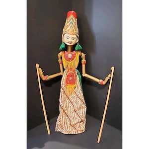May include: An Indonesian wooden puppet with a detailed headdress and facial features. The puppet has a red and gold bodice, a patterned skirt, and green accents. It holds two wooden control rods. The puppet's height is approximately 40 cm.